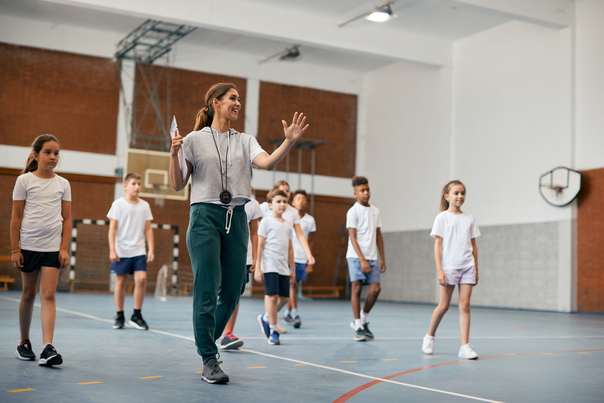 Young female coach having PE class with group of elementary students at school gym.